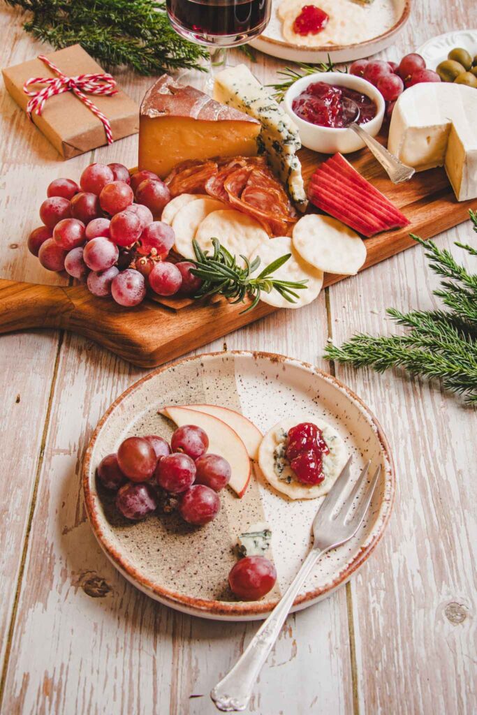 Tábua de queijos natalina com queijos variados, frutas frescas, geleia, crackers e ervas, com elementos decorativos natalinos ao fundo.