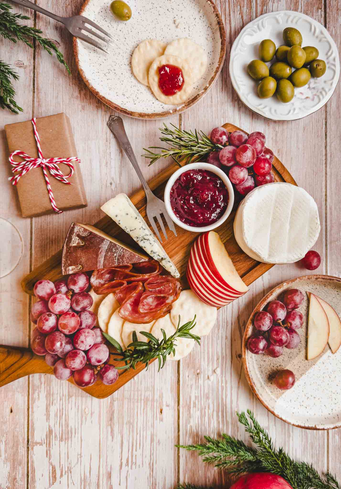 Tábua de queijos decorada com uvas, maçãs, alecrim, embutidos e uma pequena tigela de geleia vermelha, compondo um cenário acolhedor e festivo.