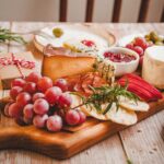 Prato com biscoitos crocantes cobertos com geleia vermelha, queijo azul e azeitonas verdes, ao lado de uma tábua de frios com queijos, frutas e embutidos.