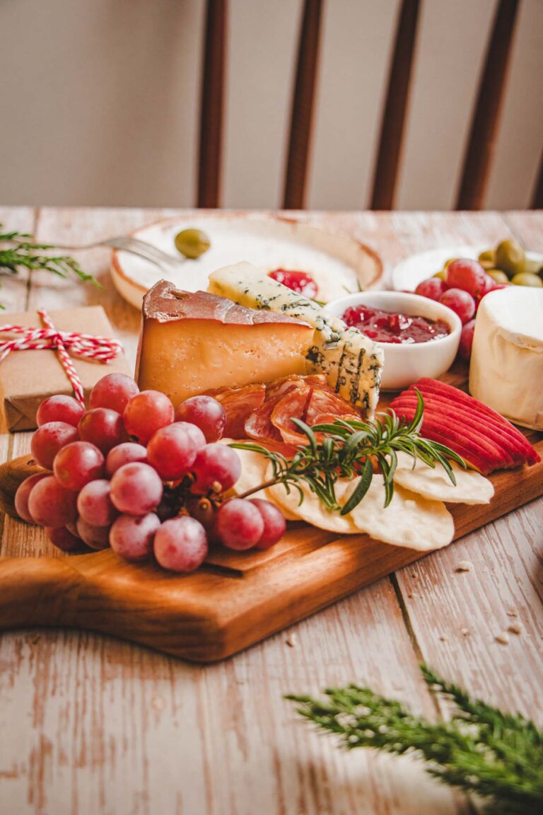 Prato com biscoitos crocantes cobertos com geleia vermelha, queijo azul e azeitonas verdes, ao lado de uma tábua de frios com queijos, frutas e embutidos.