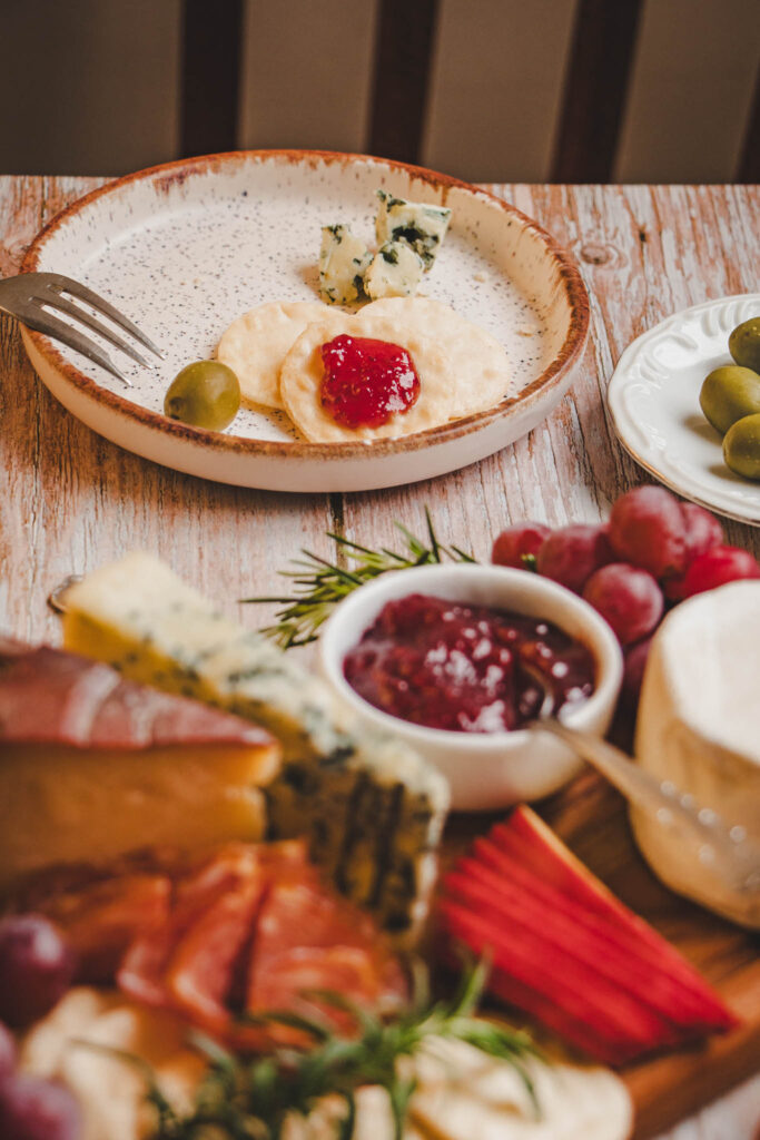 Prato com biscoitos crocantes cobertos com geleia vermelha, queijo azul e azeitonas verdes, ao lado de uma tábua de frios com queijos, frutas e embutidos.